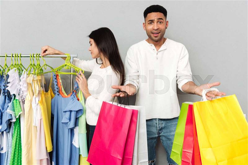 Image of young confused man holding shopping bags while his woman choosing clothes over grey wall, stock photo
