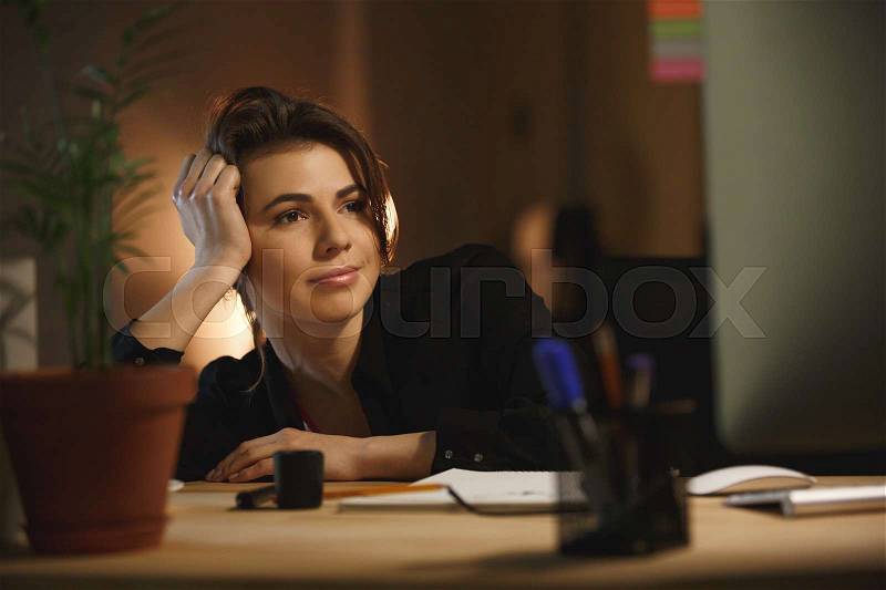 Picture of concentrated young woman designer sitting in office at night using computer. Looking aside, stock photo