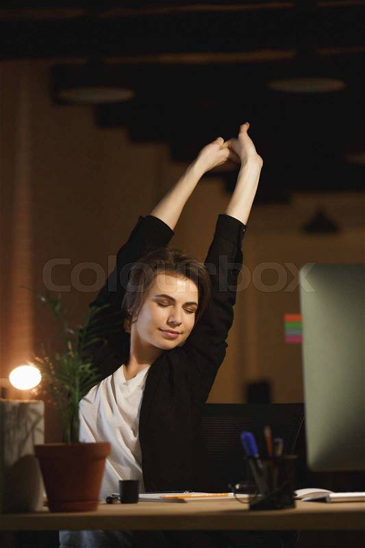 Image of tired young woman designer sitting in office at night using computer while stretching, stock photo
