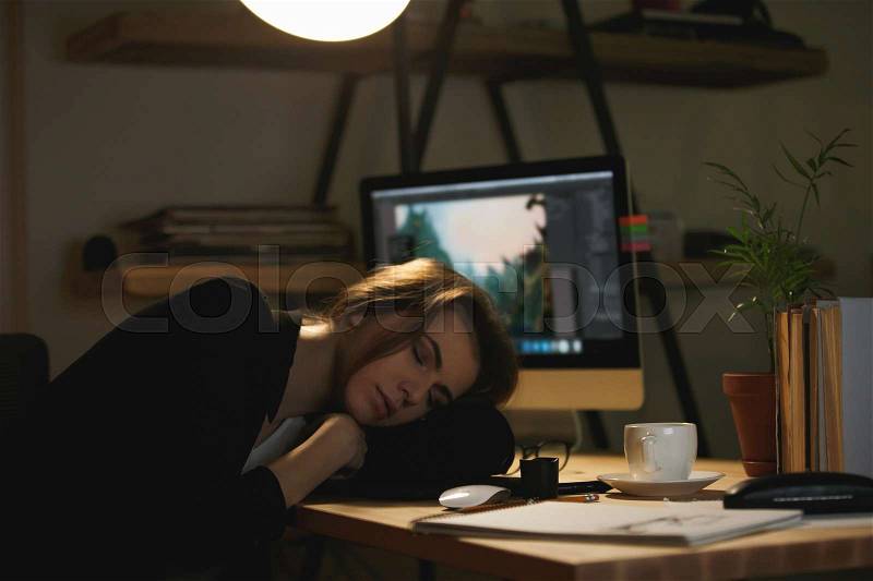 Image of young lady designer sleeping on workspace indoors at night near computer and graphics tablet, stock photo