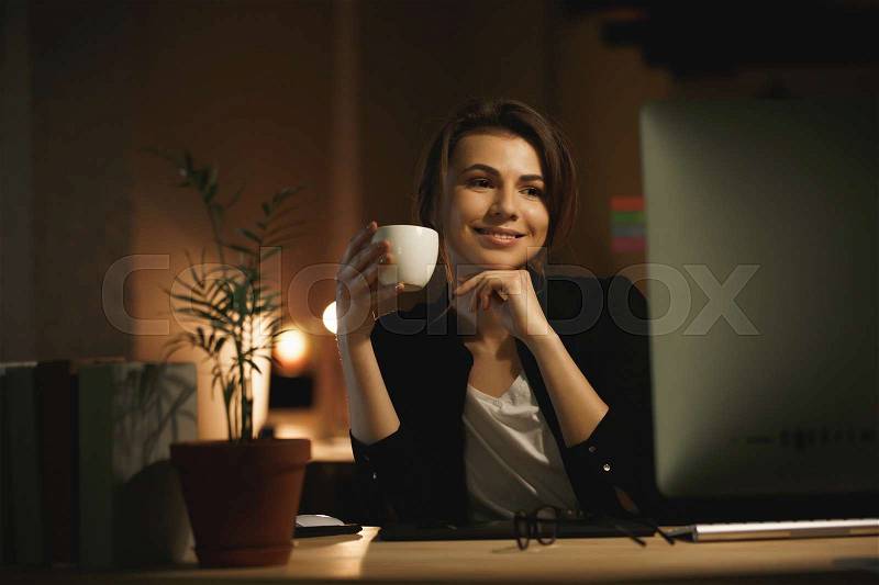 Photo of happy young woman designer sitting indoors at night using computer and drinking coffee. Looking aside, stock photo