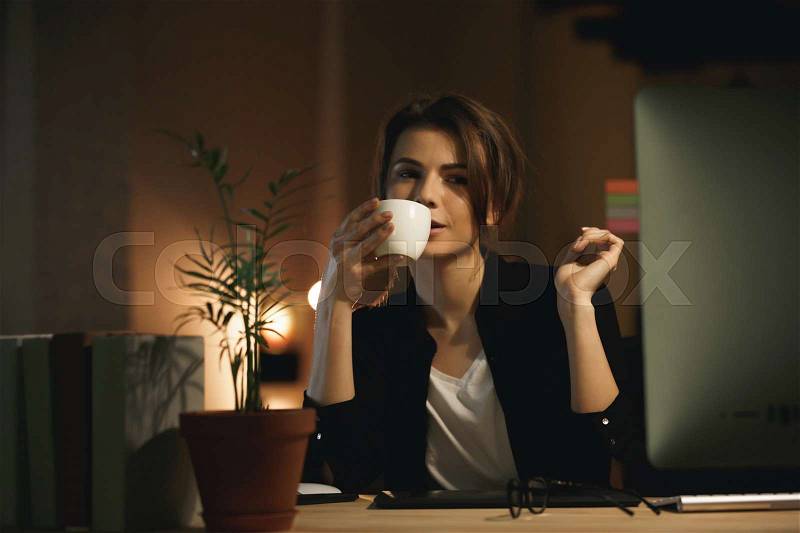 Image of concentrated young woman designer sitting indoors at night using computer and drinking coffee. Looking aside, stock photo
