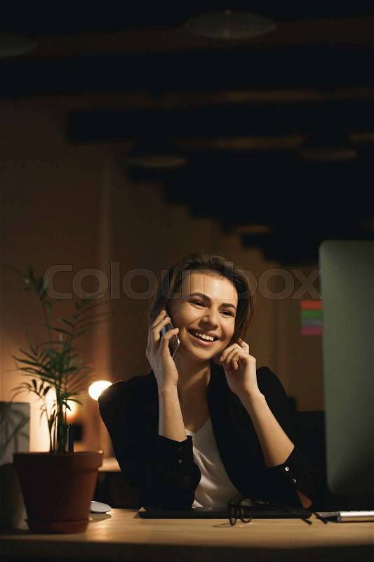 Image of smiling young woman designer sitting indoors at night using computer and talking by phone. Looking aside, stock photo