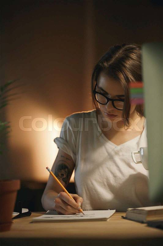 Picture of serious young woman designer sitting indoors at night writing notes using computer. Looking aside, stock photo