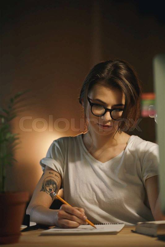 Image of concentrated young woman designer sitting indoors at night writing notes using computer. Looking aside, stock photo