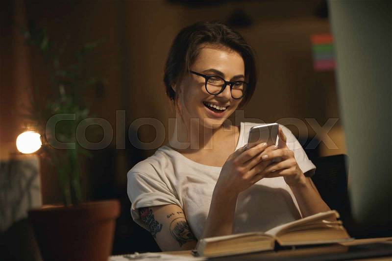 Image of young happy lady designer sitting indoors at night chatting by mobile phone. Looking aside, stock photo