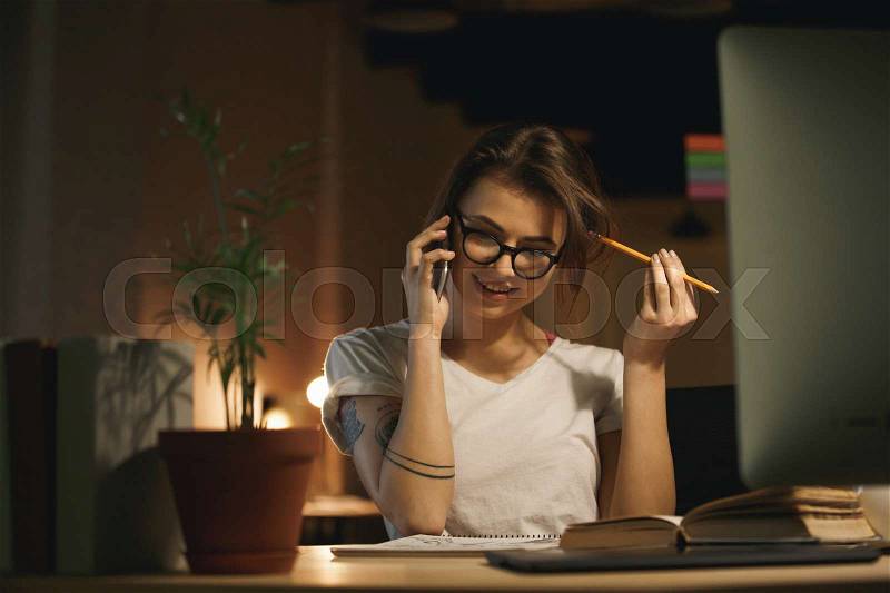 Image of young concentrated woman designer sitting indoors at night talking by mobile phone and holding pencil. Looking aside, stock photo