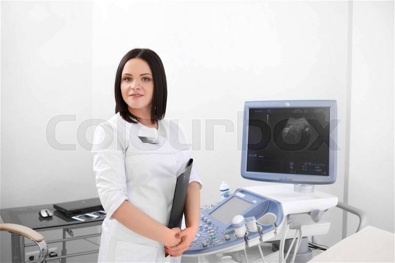 Pretty brunette female doctor against ultrasound equipment and computer, waiting for patients. Professional nurse in white uniform smiling at camera in cabinet at modern hospital. Medicine concept, stock photo
