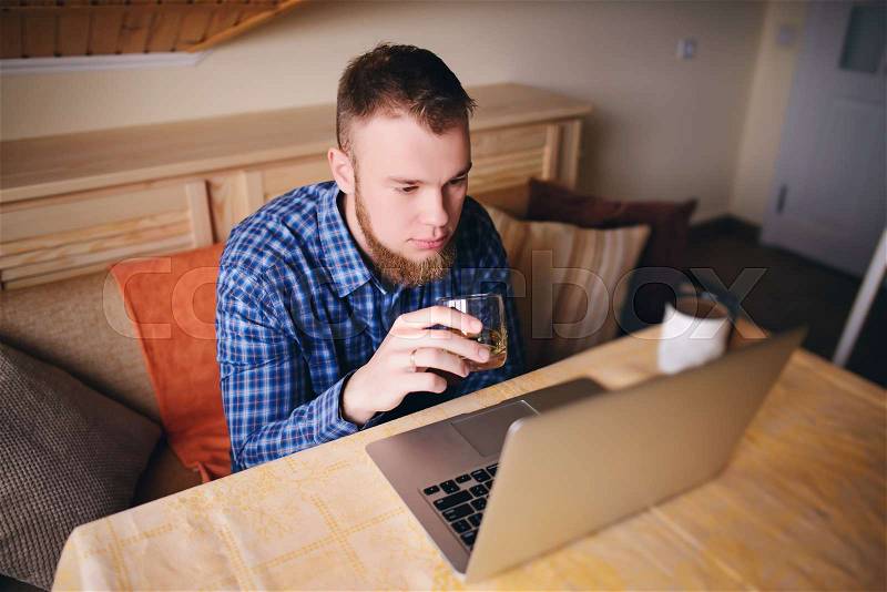 Young professional surfing the Internet on his laptop and drinking whiskey from a glass, stock photo