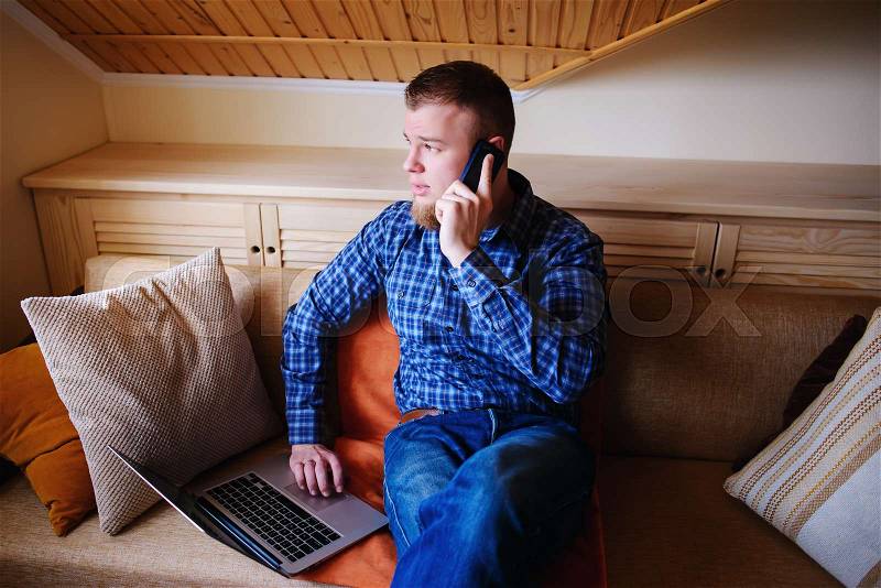 Young man using his smartphone for online banking - sitting on sofa with laptop on leap, stock photo