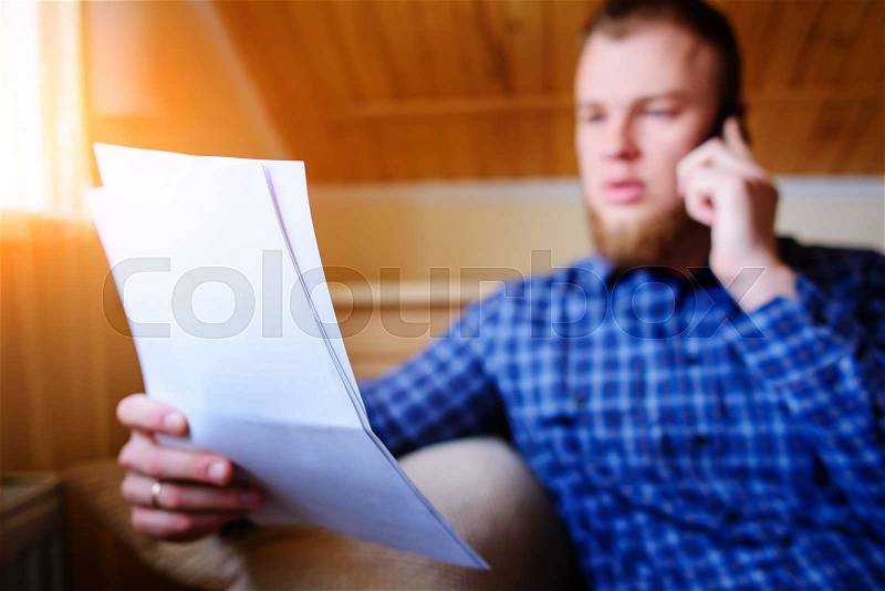 Discussing that document. Serious mature man holding paper and talking on the mobile phone while sitting on the couch at home, stock photo