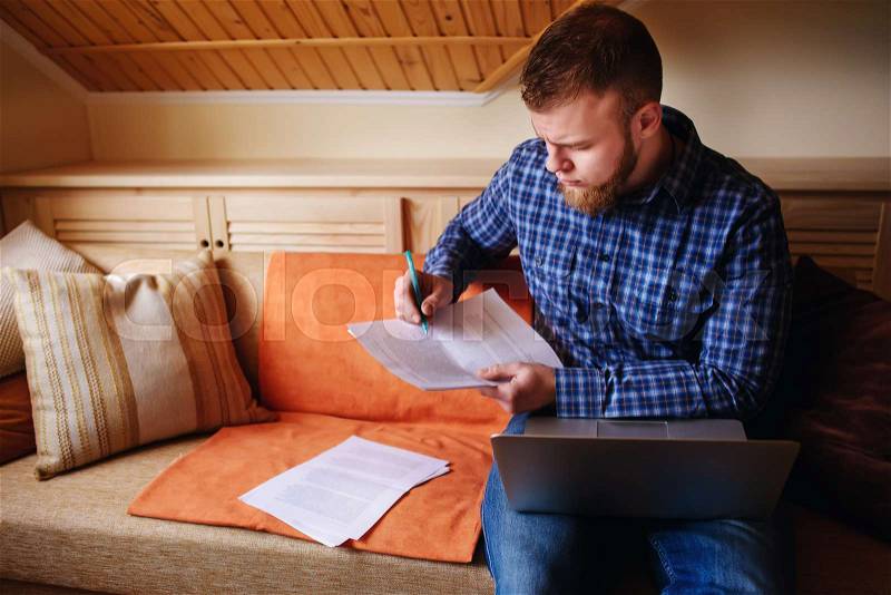 Young man studying from home sitting on a sofa holding documents while working on a laptop computer, stock photo