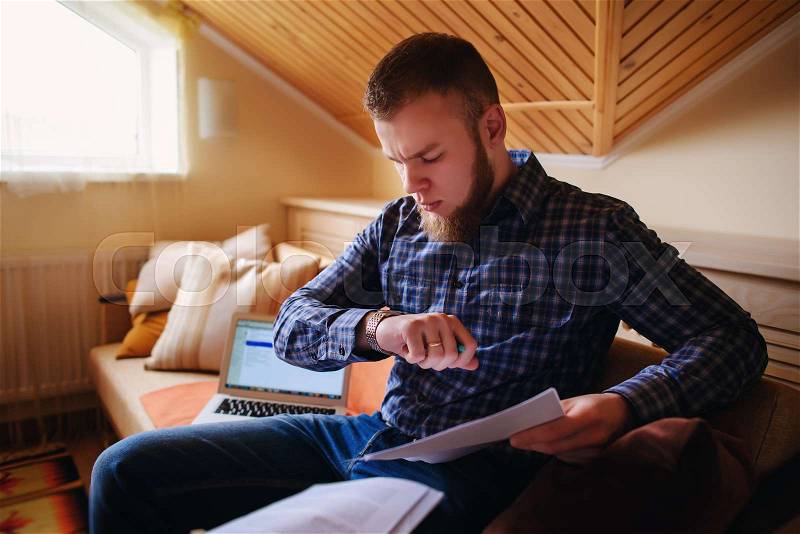 Young man studying from home sitting on a sofa holding documents while working on a laptop computer, stock photo