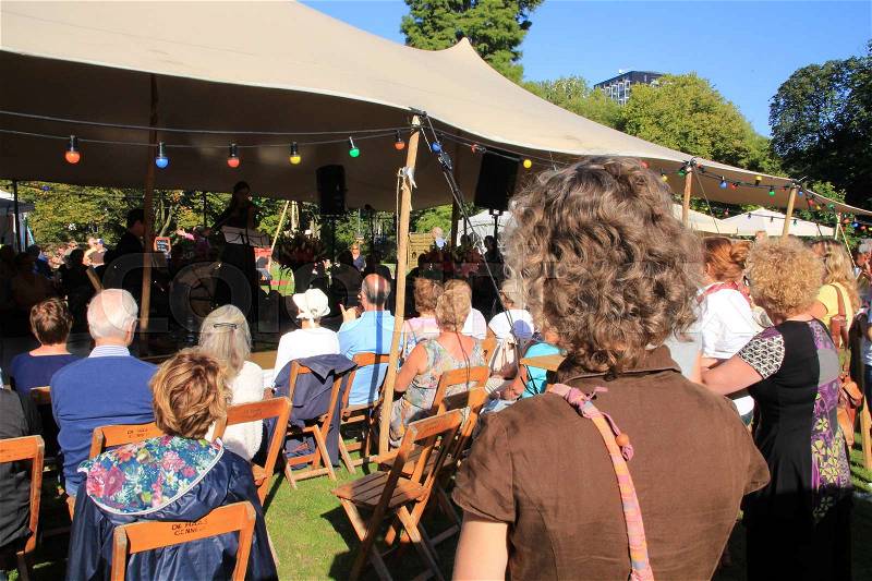 People, standing or sitting on the folding chairs listening to the music uner the tent during the event in the urban park in the summer, stock photo