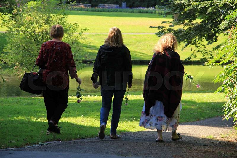 Three ladies are walking with a pink rose in their hands in the shadow in the urban park in the summer, stock photo