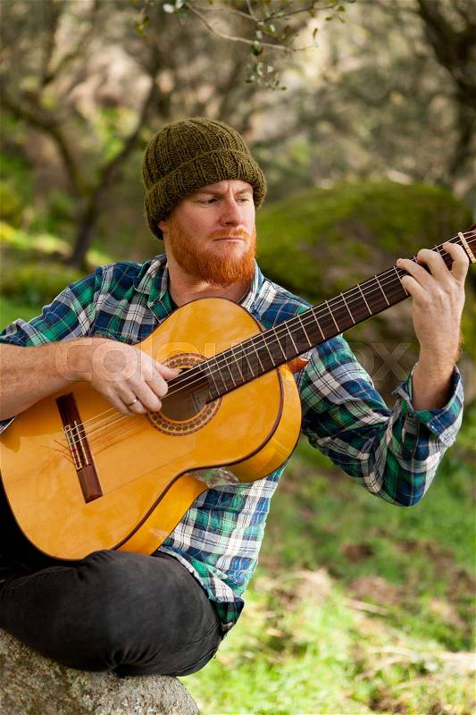Hipster man with red beard playing a guitar in the field, stock photo