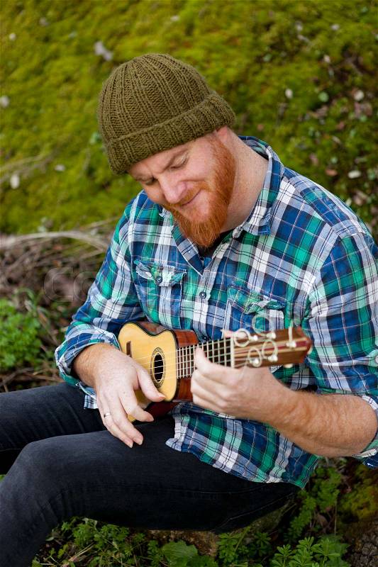 Hipster man with red beard playing a ukulele in the field, stock photo