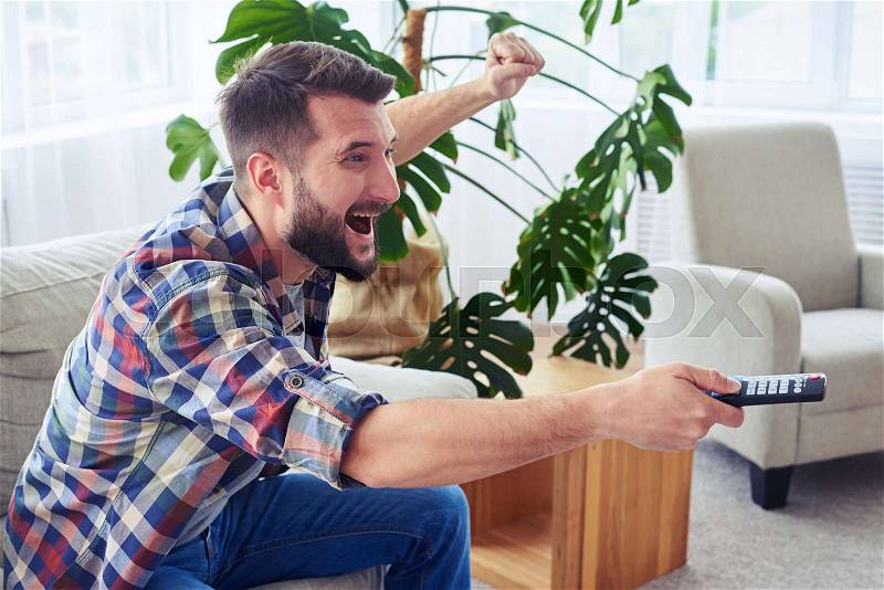 Mid shot of attractive man cheering and switching channel with remote control, stock photo
