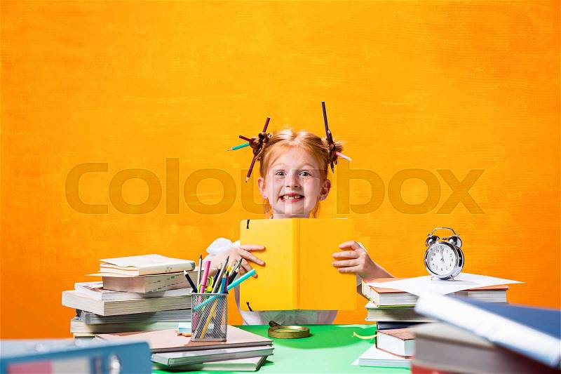 The Redhead teen girl with lot of books sitting with notebook at table on orange studio background. The education and back to school concept, stock photo