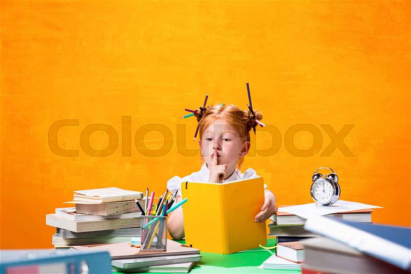 The Redhead teen girl with lot of books sitting at table on orange studio background. The education and back to school concept, stock photo