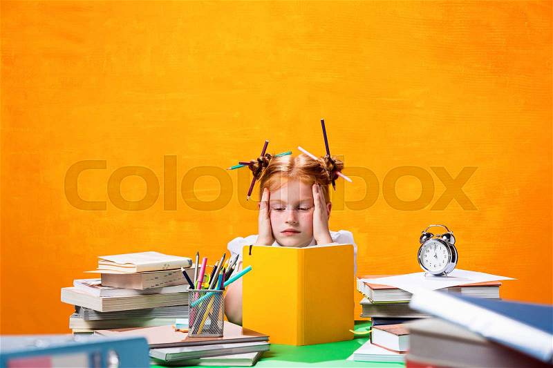 The Redhead teen girl with lot of books sitting at table on orange studio background. The education and back to school concept, stock photo