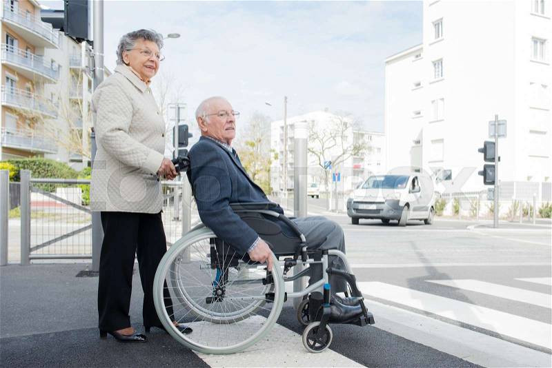 Elderly lady pushing husband in wheelchair across road, stock photo