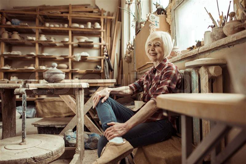 Happy senior woman sitting on bench near window at workshop, stock photo