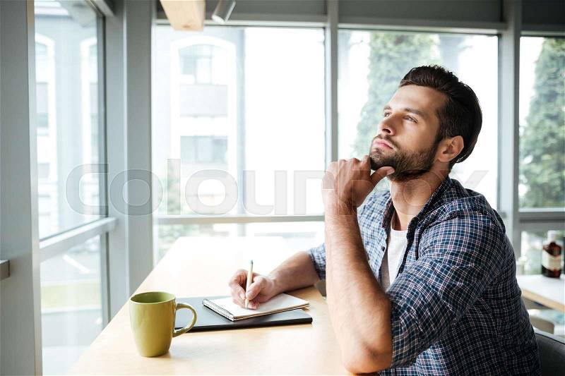 Image of young thinking man in office coworking while writing notes drinking tea. Looking aside, stock photo