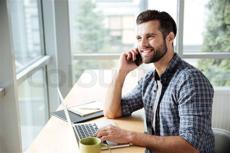 Photo of young happy man in office coworking while using laptop computer. Looking aside while talking by phone, stock photo