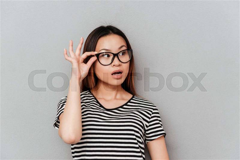 Shocked asian woman looking away and holding her glasses isolated over grey, stock photo