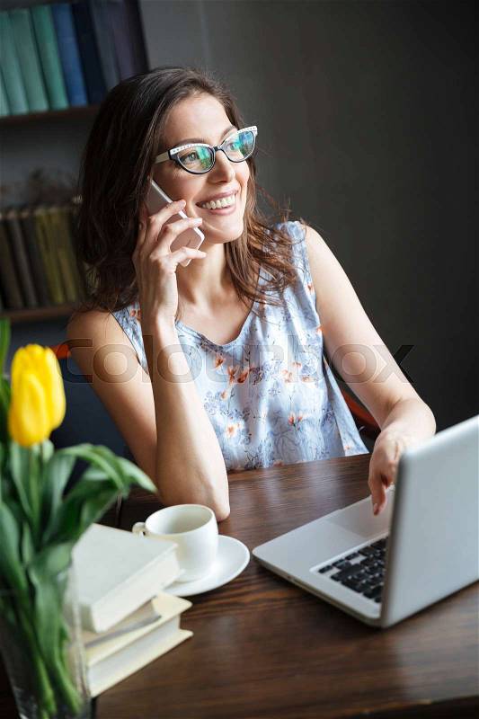 Image of beautiful woman writer sitting indoors while talking by phone and using laptop computer. Looking aside, stock photo