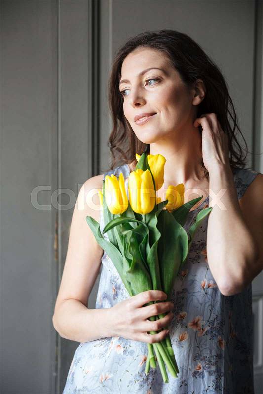 Image of happy woman writer sitting indoors while holding tulips. Looking aside, stock photo
