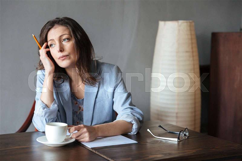 Image of thinking serious woman writer sitting indoors while drinking coffee and writing notes. Looking aside, stock photo