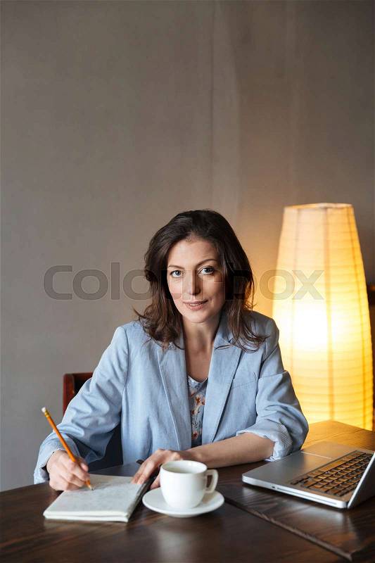 Picture of thinking woman writer sitting indoors using laptop computer. Looking at camera writing notes, stock photo