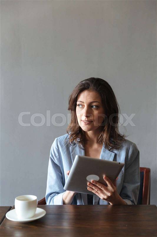 Picture of amazing woman sitting indoors. Looking aside using tablet computer, stock photo