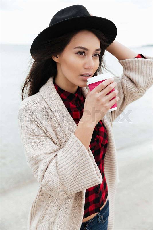 Image of pretty young caucasian lady walking outdoors at beach wearing warm jacket drinking coffee. Looking aside, stock photo