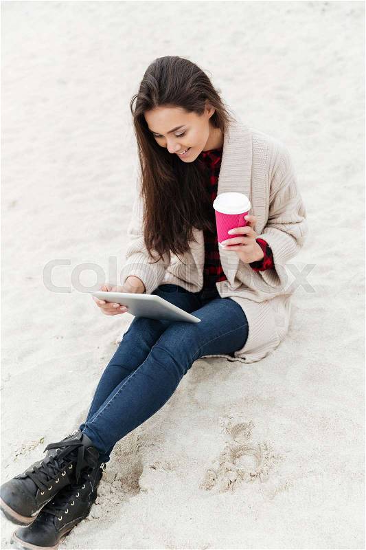 Image of young caucasian lady sitting outdoors at beach wearing warm jacket using tablet computer. Looking aside and drinking coffee, stock photo