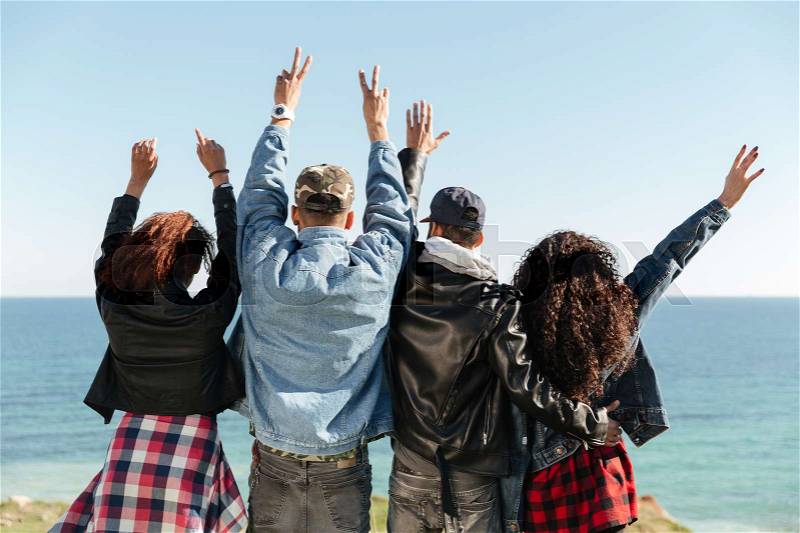 Back view image of a group of friends standing outdoors near beach with raised hands, stock photo