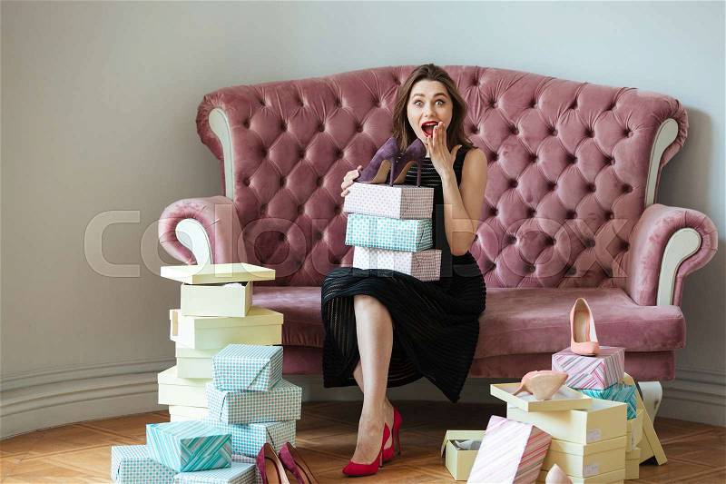 Picture of smiling young lady sitting on sofa indoors choosing shoes. Looking at camera, stock photo