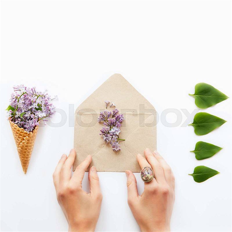 Square card with women hands touching craft paper envelope with lilac flowers surrounded by a waffle corn with a bouquet of lilac and a row of green leafs on white background. Flat lay. Text space, stock photo