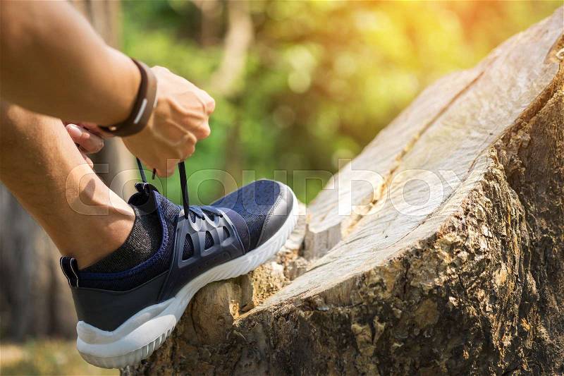 Cropped shot of young man runner tightening running shoe laces, getting ready for jogging exercise outdoors. Male jogger lacing his sneakers standing on forest path before morning run, stock photo