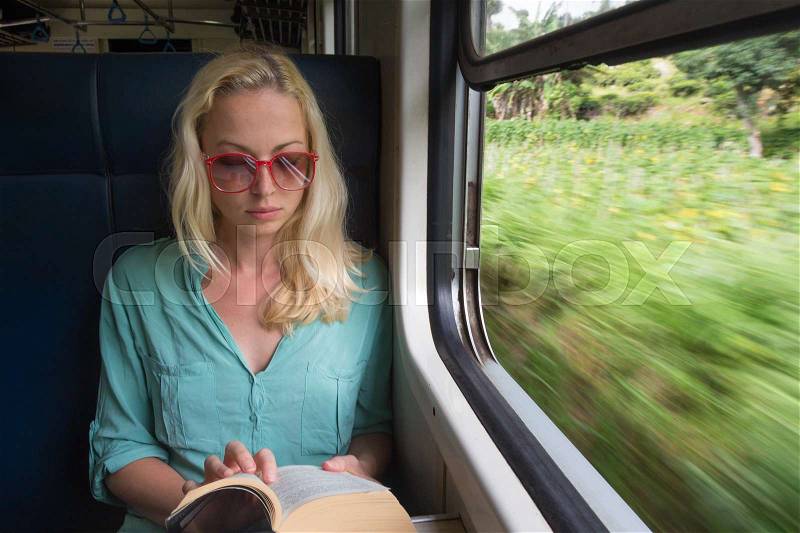 Blonde caucasian woman reading book by train window , traveling by train in third world country. Blured green fields can be seen trough window, stock photo