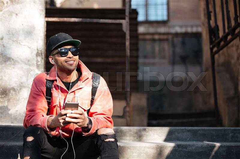 Portrait of a smiling stylish african man in leather jacket sitting on a stairs and listening to music with earphones outdoors on a street, stock photo