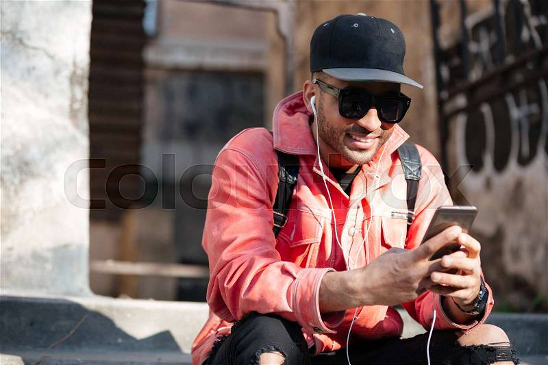Portrait of a confident stylish african man in leather jacket sitting on a stairs and listening to music with earphones outdoors on a street, stock photo
