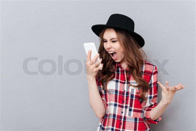 Image of screaming angry young woman standing over grey wall wearing hat talking by mobile phone. Looking aside, stock photo