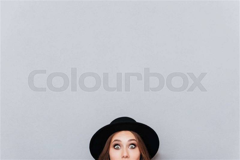 Portrait of a surprised young woman in hat peeping out from the edge and looking at camera isolated over gray background, stock photo