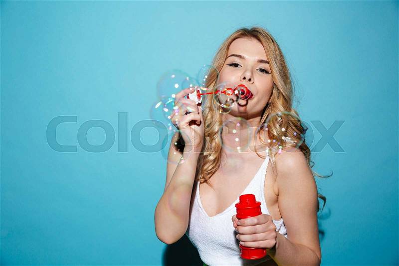 Portrait of a young attractive woman in summer clothes blowing soap bubbles isolated over blue background, stock photo