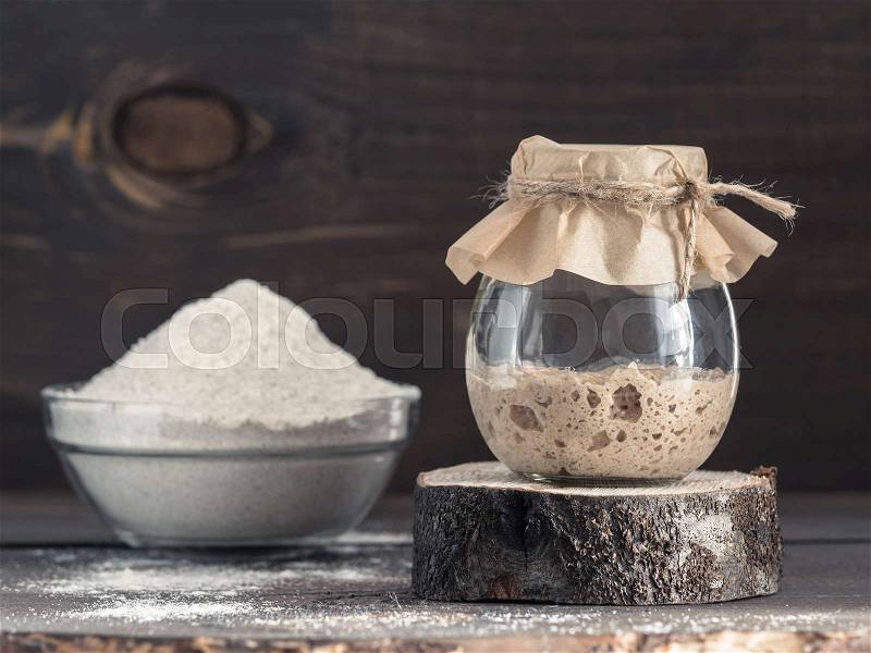 Active rye sourdough starter in glass jar and rye flour on brown wooden background. Starter for sourdough bread. Toned image. Copy space, stock photo
