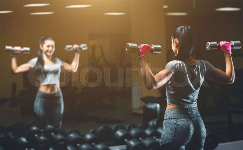 Slim, bodybuilder girl, lifts heavy dumbbell standing in front of the mirror while training in the gym. Sports concept, fat burning and a healthy lifestyle, stock photo