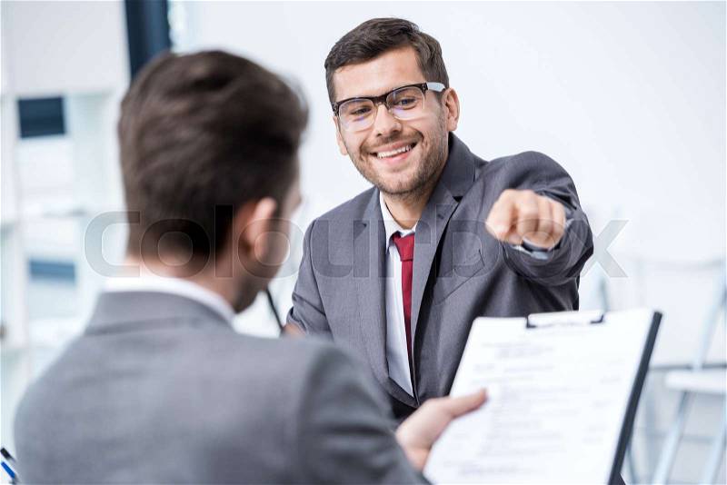 Smiling man in suit and eyeglasses pointing at clipboard at job interview, business concept, stock photo